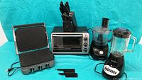 Photo of Black & Decker panini press open, toaster oven with front glass door and dials, blender with control panel, and food processor with feed chute, all placed on a surface with a turquoise background.
