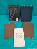 Photo showing four spiral-bound notebooks (three brown, one blue) and one shrink-wrapped black address book with gold envelope icon, all arranged on a teal background.
