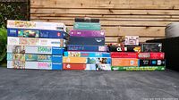 Photo showing a large stack of boxed puzzles with piece counts from 500 to 1500 pieces. Boxes have varied artwork including family scenes, landscape collages, cats, and abstract designs.
