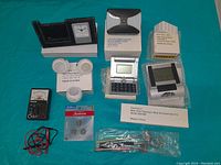 Photo showing an assortment of small electronics, lighting devices, calculator, multimeter, and accessories arranged on a teal surface.