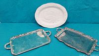 Front view of two metal trays with ornate handles and white ceramic oval tray behind them.