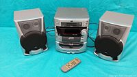 JVC three-piece stereo system with two speakers and original remote on a blue backdrop.