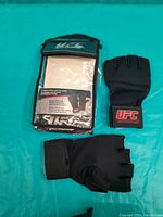 Photo shows a pair of black mixed martial arts gel gloves with UFC branding. One glove is outside the packaging, the other remains inside a transparent plastic bag with product details.