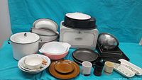 Image showing a collection of cookware including enamel-coated pots, Pyrex dishes, pans, mugs, and a metal breadbox against a turquoise background.