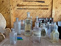 Photo shows ten antique glass bottles of various shapes and sizes on a table in a wooden paneled room.
