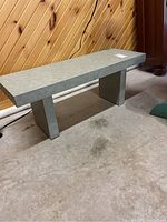 Full view of solid wood bench with green laminate surface, rectangular top and two supporting legs in a room with wood paneling and carpeted floor.