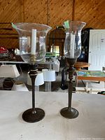 Two tall brass candle holders with large clear glass shades and white candles inside, shown on a table with wood panel background.