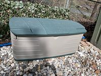 Rubbermaid outdoor deck box with green lid placed on a bed of stones outdoors.