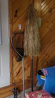 Fishing rods, large fishing net with metal handle, and tiki style patio umbrella in closed position leaning against wood panel wall.