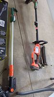 Overview of Black and Decker cordless grass trimmer/edger on right with orange handle and black attachments alongside a long extension arm with green and orange handle on left, metal shafts visible.