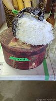 White feathery ladies hat displayed on top of a floral and fruit designed round hat box, showing the full hat and box.
