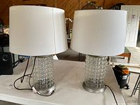 Two lamps with hollow textured glass bases and round metal bottoms each fitted with a white drum shade, shown on white table.