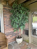 Front view of large faux ficus tree with variegated leaves inside a decorative beige ceramic pot, positioned against a brick wall on a stone patio with wicker chair nearby.