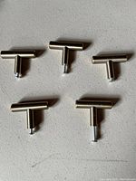 Five small T-shaped champagne gold cabinet pulls with screw bases arranged on a flat surface.