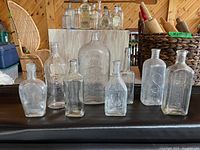 Nine vintage clear glass bottles shown on black surface against wooden background. Varied shapes, some embossed with designs, many missing labels.