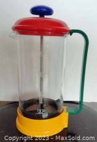 Front view of colorful Memphis style coffee pot French press with red lid, blue knob, green handle, and yellow base, made of glass and plastic or metal parts. Marked BMF inside a triangle on the glass.