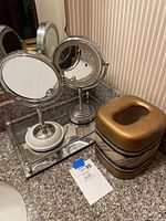 Two round tabletop magnifying mirrors with chrome finish standing on white weighted bases, placed on a rectangular mirrored tray. Ceramic tissue box cover with bronze and black design next to the tray on granite countertop.