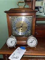 Front view of all three clocks placed on a wooden table: one large wooden mantle clock with ornate dial and two smaller silver Elgin alarm clocks.