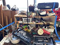 Overall view of a cluttered garage workspace showing a black Craftsman socket set open with assorted sockets, a red Craftsman branded power tool, assorted small containers, cords, and a black Sony radio cassette player on top shelf.