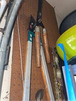 Manual hedge trimmers with wooden handles and a large blue and white handled cutting tool hanging on pegboard wall