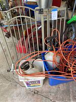 Photo of the lot showing two handheld garden sprayers with black handles in a plastic tote, several orange extension cords tangled together, and one beige metal adjustable gate leaning against a surface.