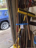 Photo showing assorted gardening tools including a broom with yellow bristles and various handles for shovels, tree trimmer, and hoe arranged upright.