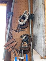 Photo showing various sizes of metal C-clamps, two wooden hand screw clamps, and a metal pipe clamp hanging on pegboard.