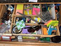 Top view of open kitchen drawer showing multiple compartments containing sunglasses, Post-it notes in different colors, a black handled knife, and miscellaneous stationery items.