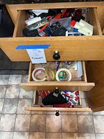 Photo of three wooden drawers partially open showing assorted household items inside including flashlights, tapes in rolls, dish towels, and various small tools and electronics.
