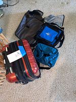 Photo of all four items together including red hard shell rolling suitcase and three fabric tote bags in black, blue, and teal colors
