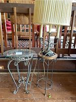 Glass top metal end table, metal plant stand with wooden top, and metal vintage-style lamp with pleated shade and marble base, placed on wooden floor against wooden furniture.