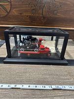 Photo of red diecast tow truck model inside black-framed glass display case with measuring tape in front showing size.