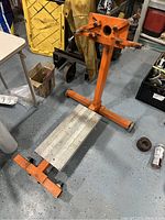 Full view of the orange metal engine stand including base, mounting arms, and rotation handle on a concrete floor.