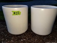 Two white ceramic planter pots side by side outdoors on mulch, showing cylindrical shape and weathering.