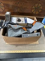 View of the box with visible items including handheld flashlight, decorative metal plaque, electronic component, and a gray bag.