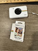 Front top view of the white Polaroid Snap camera alongside twin-pack Polaroid Premium ZINK Paper in original packaging showing packaging, camera lens, and rainbow stripe.