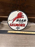 Front view of the round porcelain Gilmore gas sign showing red lion, text, and checkered flag, with a ruler for scale.