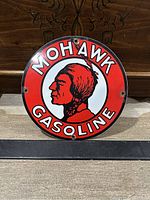 Front view of round Mohawk Gasoline porcelain sign with red background, white center, and Native American profile logo. Shows mounting holes and some chipping.