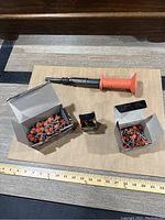 Photo showing the Remington tool with orange handle in center, two open boxes of orange-tipped powder fasteners and a small box of metal fasteners on a wooden surface.