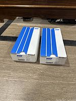 Two closed boxes of Norton sandpaper sheets, white and blue packaging, on wooden floor.