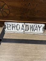 Front view of vintage Broadway street sign showing raised black letters on weathered white background with measuring tape at base indicating length.