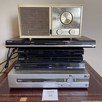 Stacked view of Zenith radio on top of five DVD players including Toshiba, Sony (two units), and Philips (two units) on wooden surface.