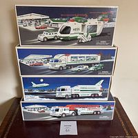 Stacked boxes of Hess toy vehicles showing detailed box art and names: Helicopter with Motorcycle and Cruiser, Toy Truck and Racers, Space Shuttle Truck and Speeders, Fire Truck.