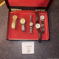 Five men's wristwatches arranged in red-lined black case, showing different band materials and watch face styles.