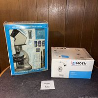 Photo of two boxed items side by side on a table: a microscope kit in a blue box with test tubes and accessories visible, and a white and blue Moen Adler faucet set box.