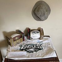 Lot arranged on table - showing Eagles XL T-shirt with many autographs, two footballs in boxes, and two caps hung on wall above