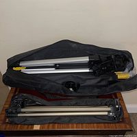 Photo showing both tripods, one inside a black carrying bag with yellow accents, the other partially outside a black carrying bag, on a carpeted floor.