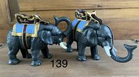 Two vintage cast iron mechanical elephant figurines side by side showing front and side views with raised trunks and detailed saddles