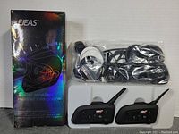 Photo of two black Ejeas V6 Pro Bluetooth helmet intercom units with buttons and antenna, packaged wired earphones and microphones, and the original box with holographic helmet art.