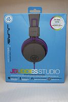 Box front of JLab JBuddies Studio headphones in purple and gray, highlighting kid-friendly features and volume limiter.